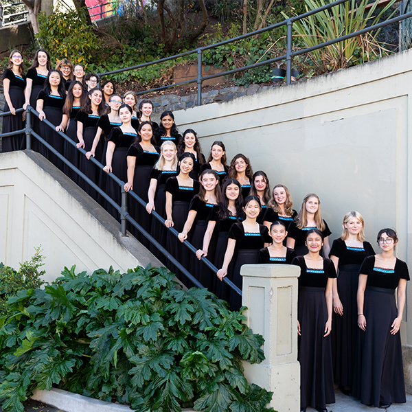 A group photo of the Young Womans Chorus wearing black dresses, standing on stairs, outside.